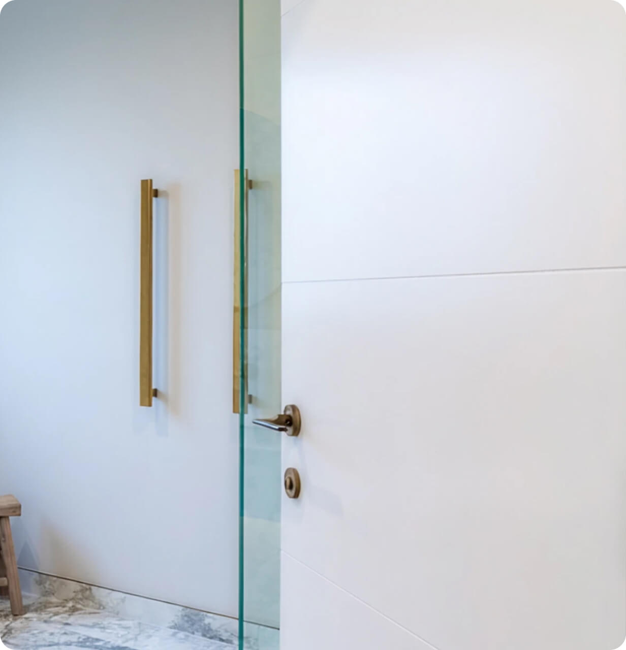 A modern, minimalistic bathroom features a glass door with brass handles and a marble floor. 