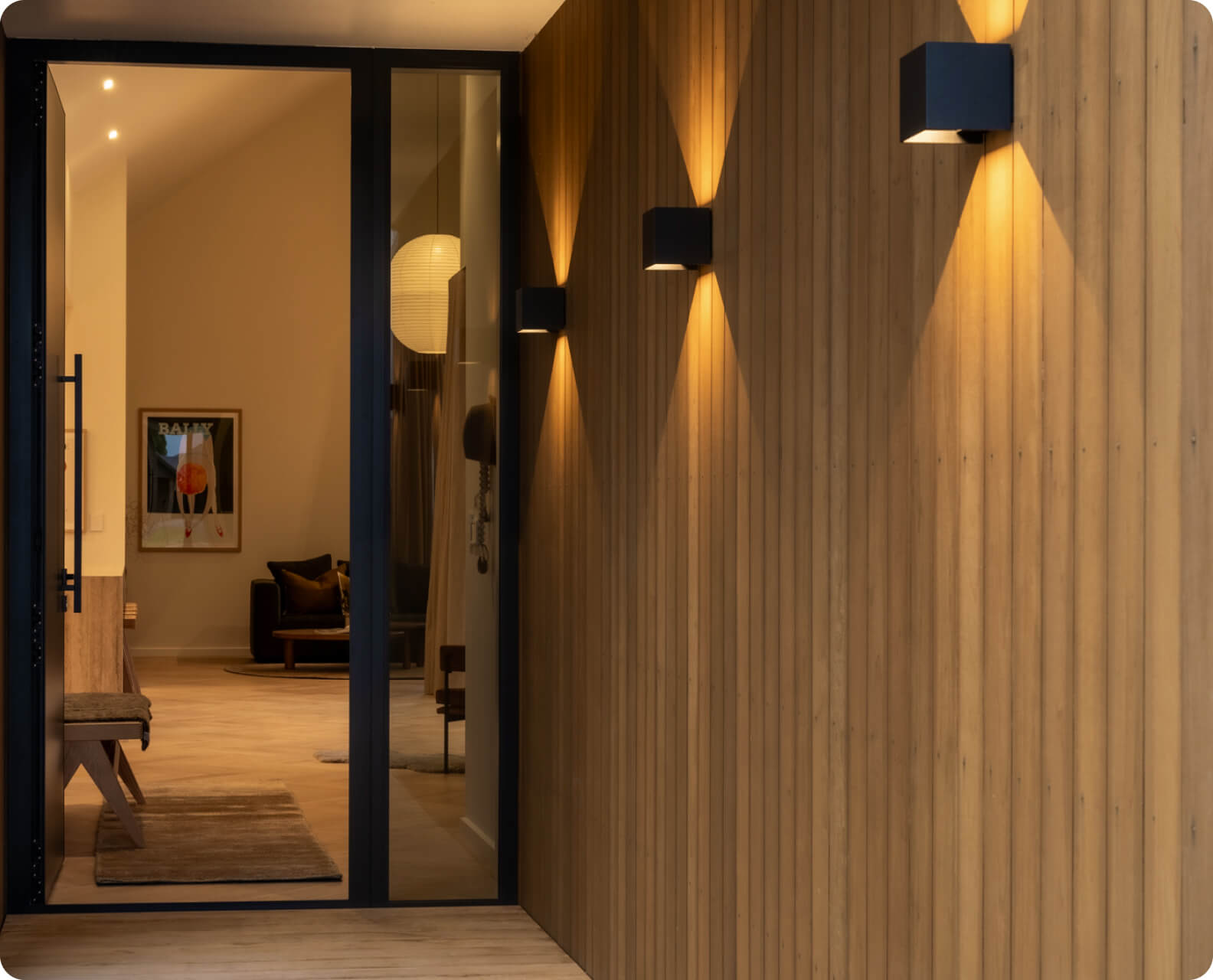 Modern interior hallway with warm wood-paneled walls, soft lighting from black wall sconces, and a glass door leading to a cozy room with artwork.