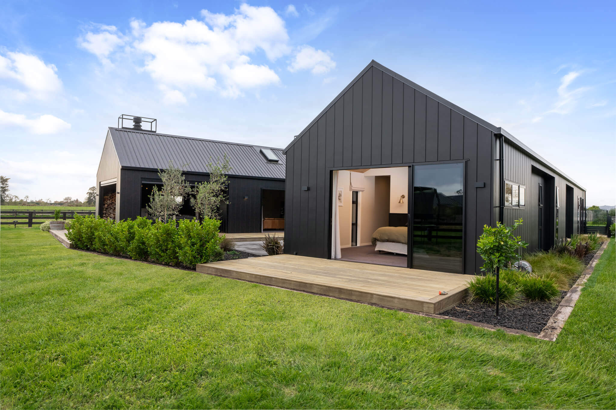 Modern black barn-style house with large windows, set amidst a lush green lawn and blue sky. 