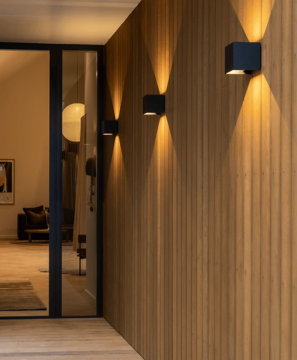 Modern entryway with wooden panel walls and soft lighting. Glass door reveals an interior with warm tones, a cozy chair, and artwork on the wall.