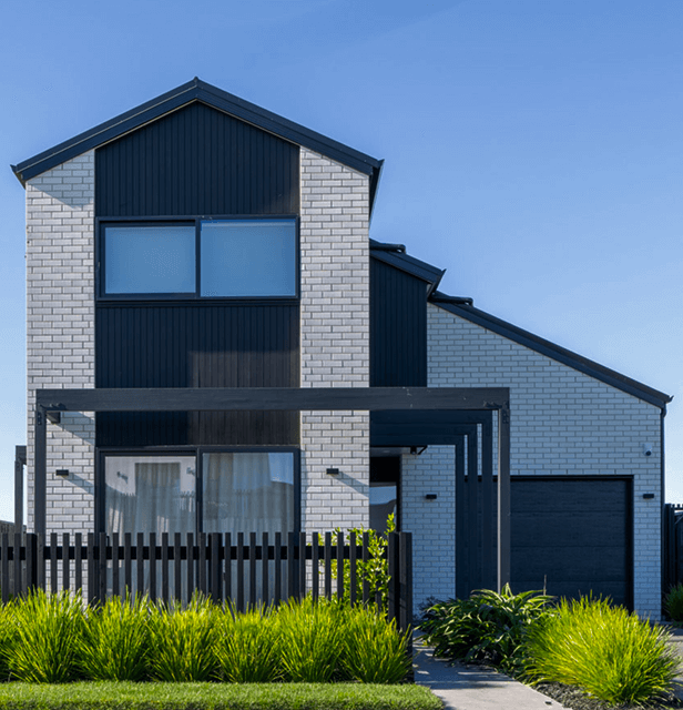 Modern two-story house with white brick and black accents, features large windows, a wooden pergola, and lush green landscaping under a clear blue sky.