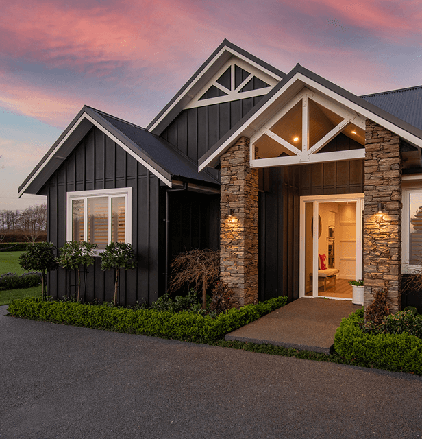 Cozy house exterior at sunset with dark siding, stone columns, and white trim. Warm lights glow from inside, surrounded by lush greenery.