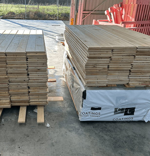 Stacks of wooden planks are neatly arranged on pallets in a warehouse, lit by natural light. 