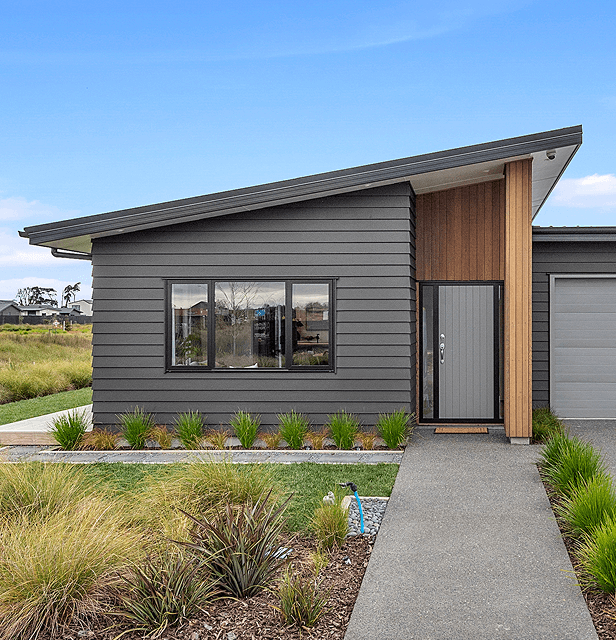 Modern single-story house with dark gray horizontal siding, large window, and slanted roof. A path leads to the entrance, surrounded by neat landscaping.