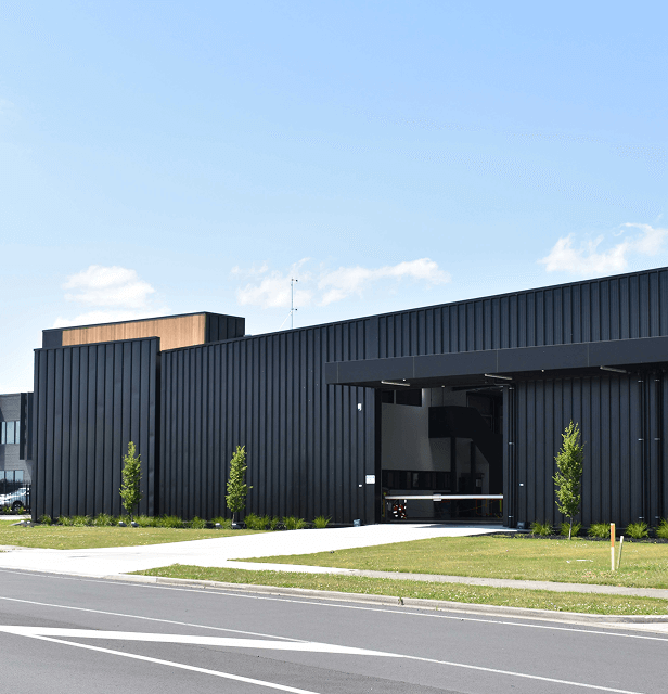 A modern industrial building with sleek black metal siding, surrounded by neatly trimmed grass and young trees under a clear blue sky with scattered clouds.
