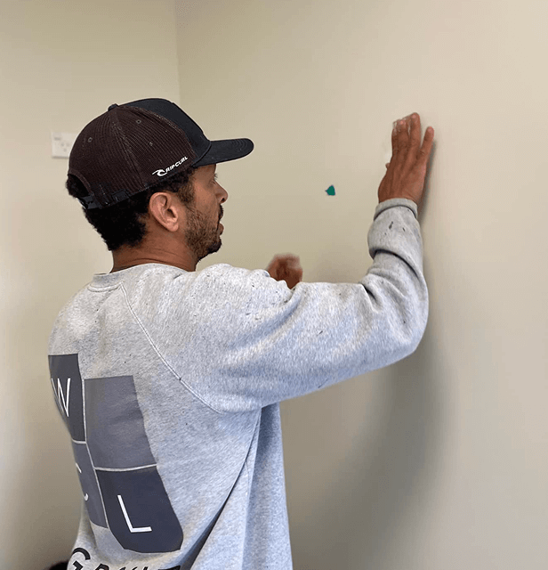 A man in a cap and gray sweatshirt stands focused, placing a small blue sticker on a light-colored wall.