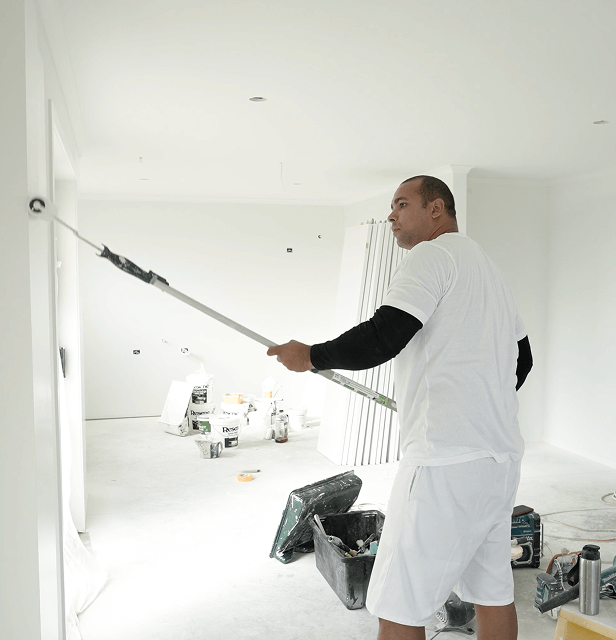 Man painting a white wall in a bright room with a paint roller extension. 