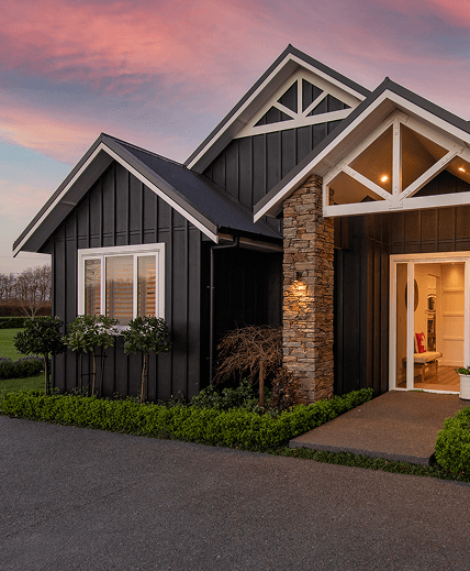 A modern house with dark siding and stone pillars is beautifully lit at dusk. 