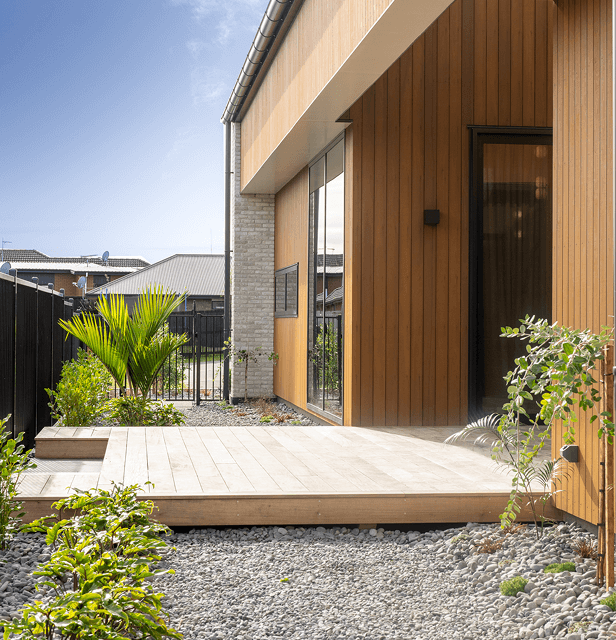 Modern patio with light wood deck, flanked by gravel and lush plants. 