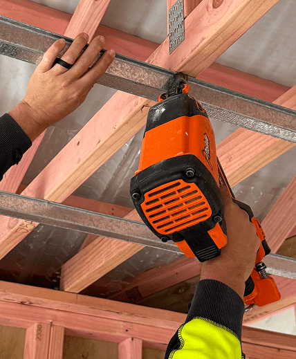A person uses an orange nail gun to install metal ceiling battens on wooden beams.