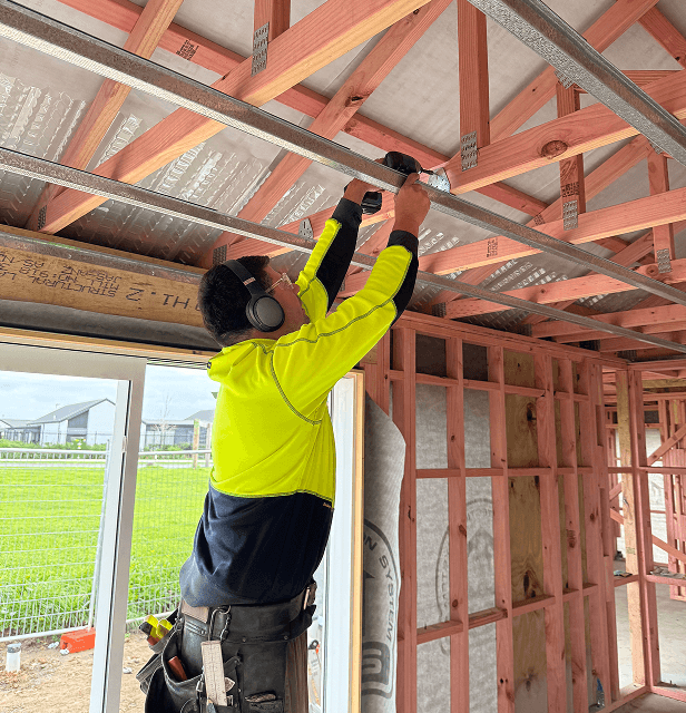 A construction worker in a neon yellow jacket uses a drill while installing metal beams in a house under construction.