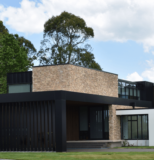 Modern house with a stone facade and black accents, large windows, surrounded by greenery and a clear blue sky.