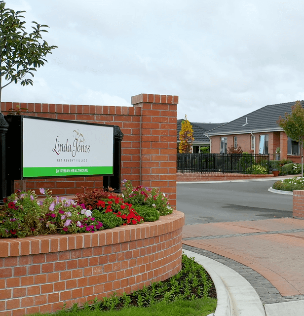 A brick entrance sign for a retirement village stands amid colorful flowers. 