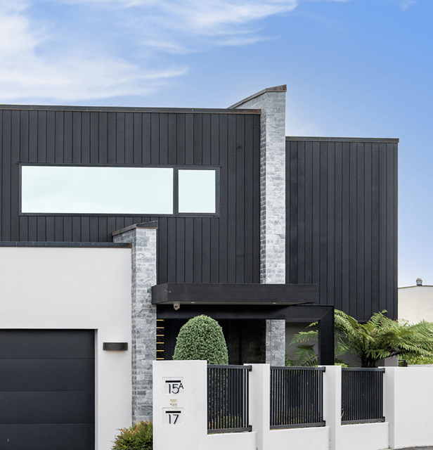 Modern house with a sleek design, featuring dark vertical panels, large horizontal window, and a white fence with plants. 