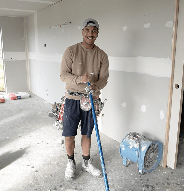 A smiling man in work clothes stands in a room under construction, holding a paint pole. 