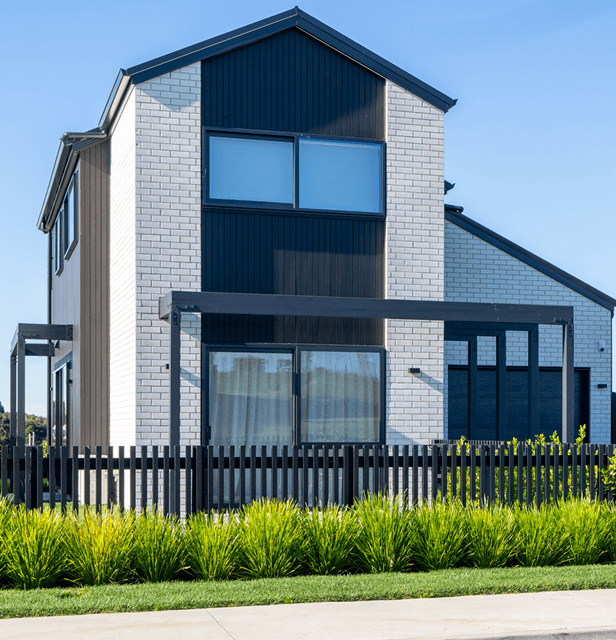 Modern two-story house with white brick and black accents, features large windows, a wooden pergola, and lush green landscaping under a clear blue sky.