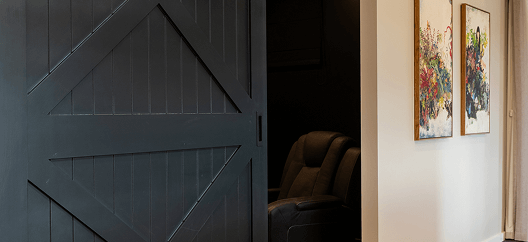 A dark sliding barn door partially reveals a cozy armchair in a dimly lit room. 
