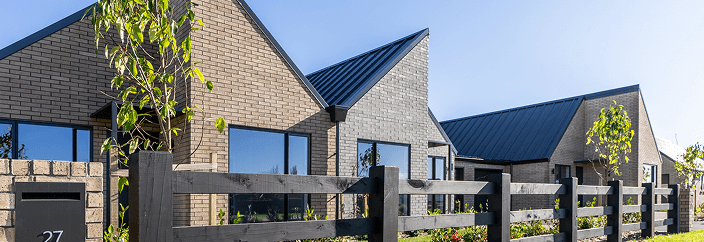 Modern townhouses with pitched roofs and brick exteriors under a clear blue sky. A black wooden fence and young trees add to the neat suburban feel.