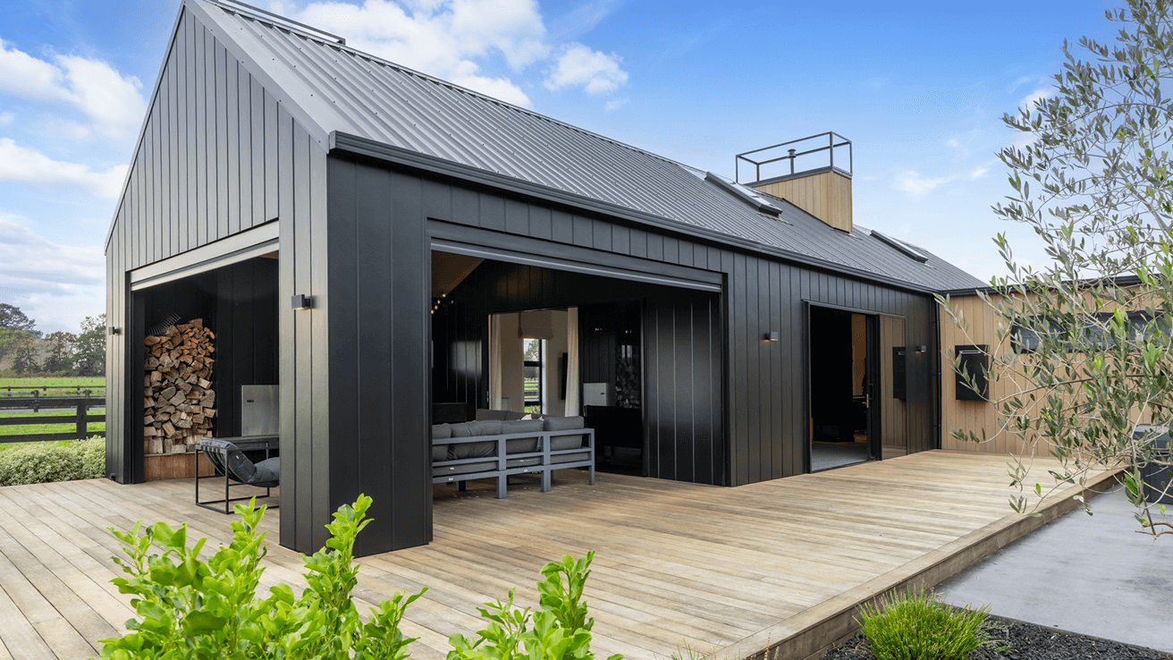 Modern black metal-clad house with wooden deck, outdoor seating, and stacked firewood. Surrounded by greenery under a bright blue sky.