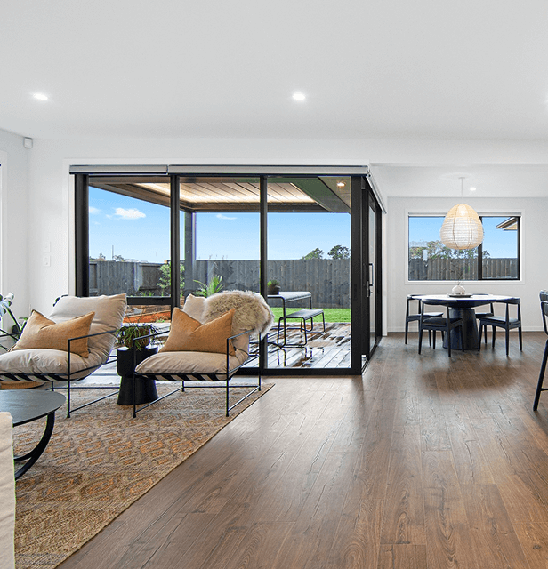 Modern open living space with hardwood floors, featuring cozy cream chairs and a rug near large glass doors. Outside, a green patio is visible.