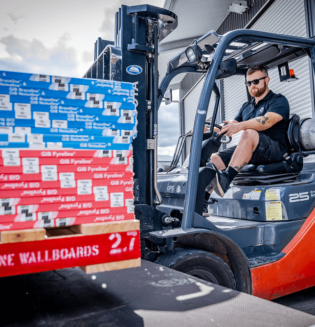 A man operates a forklift, moving stacks of colorful wallboard boxes in an industrial setting.