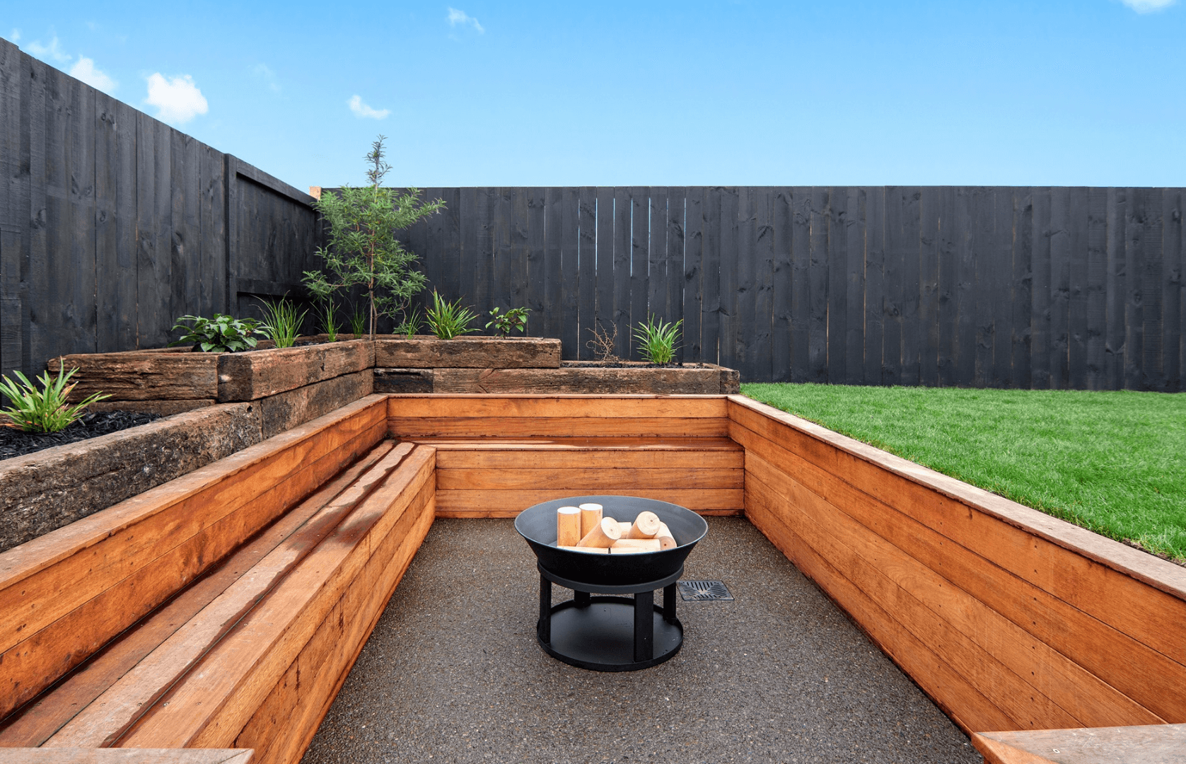 Outdoor patio with wooden seating and a central fire pit, surrounded by wooden planters and a grassy area. A tall, dark fence encloses the space.