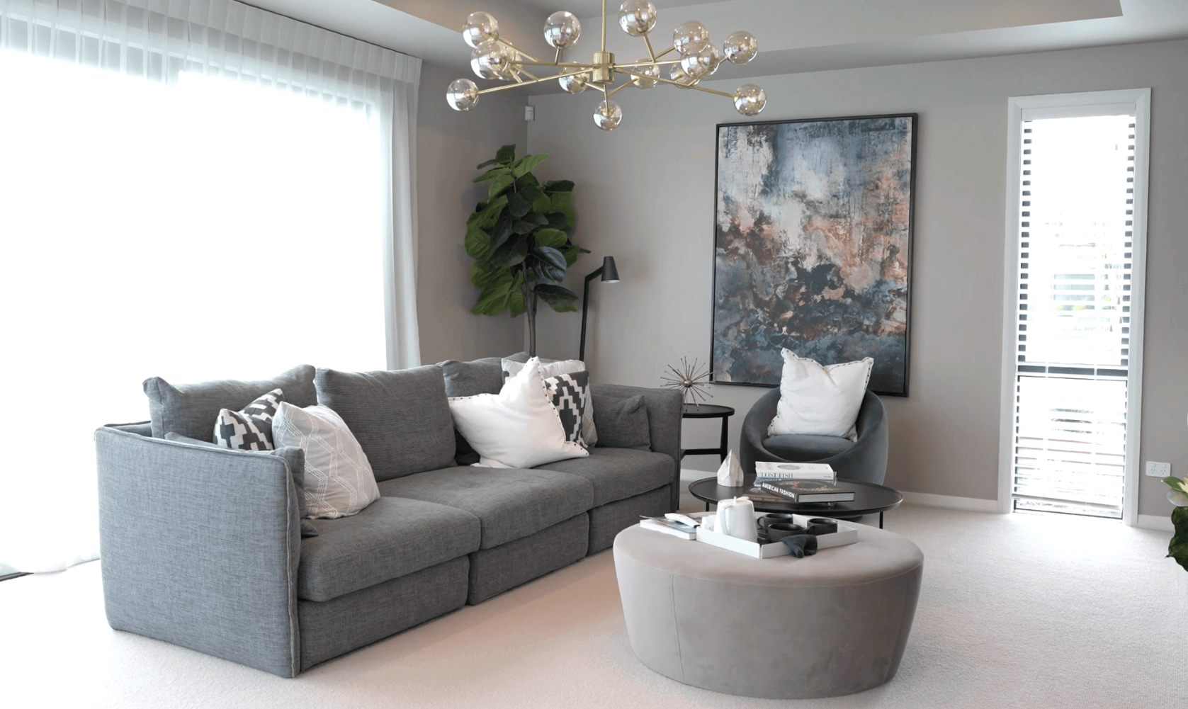 A modern living room features a gray sectional sofa with white pillows, an abstract painting, a plush chair, and a round coffee table. 