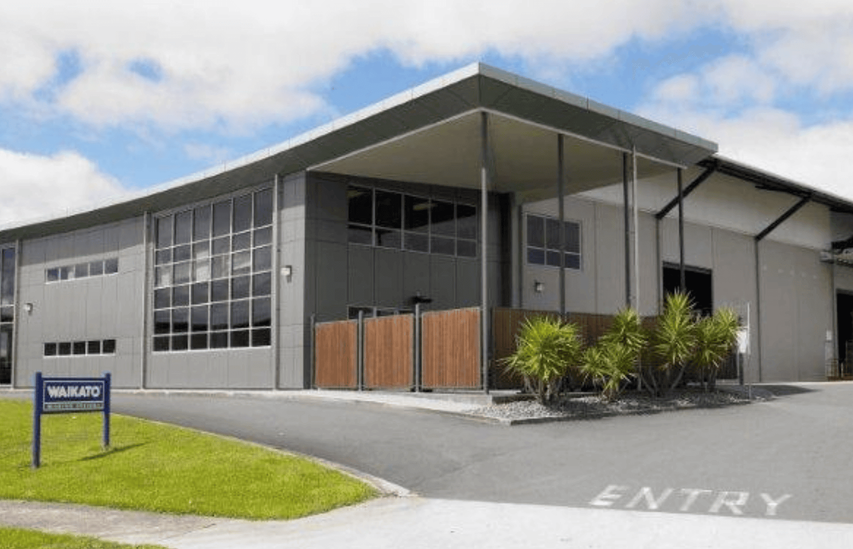 Modern industrial building with a slanted roof, large windows, and a small landscaped garden.
