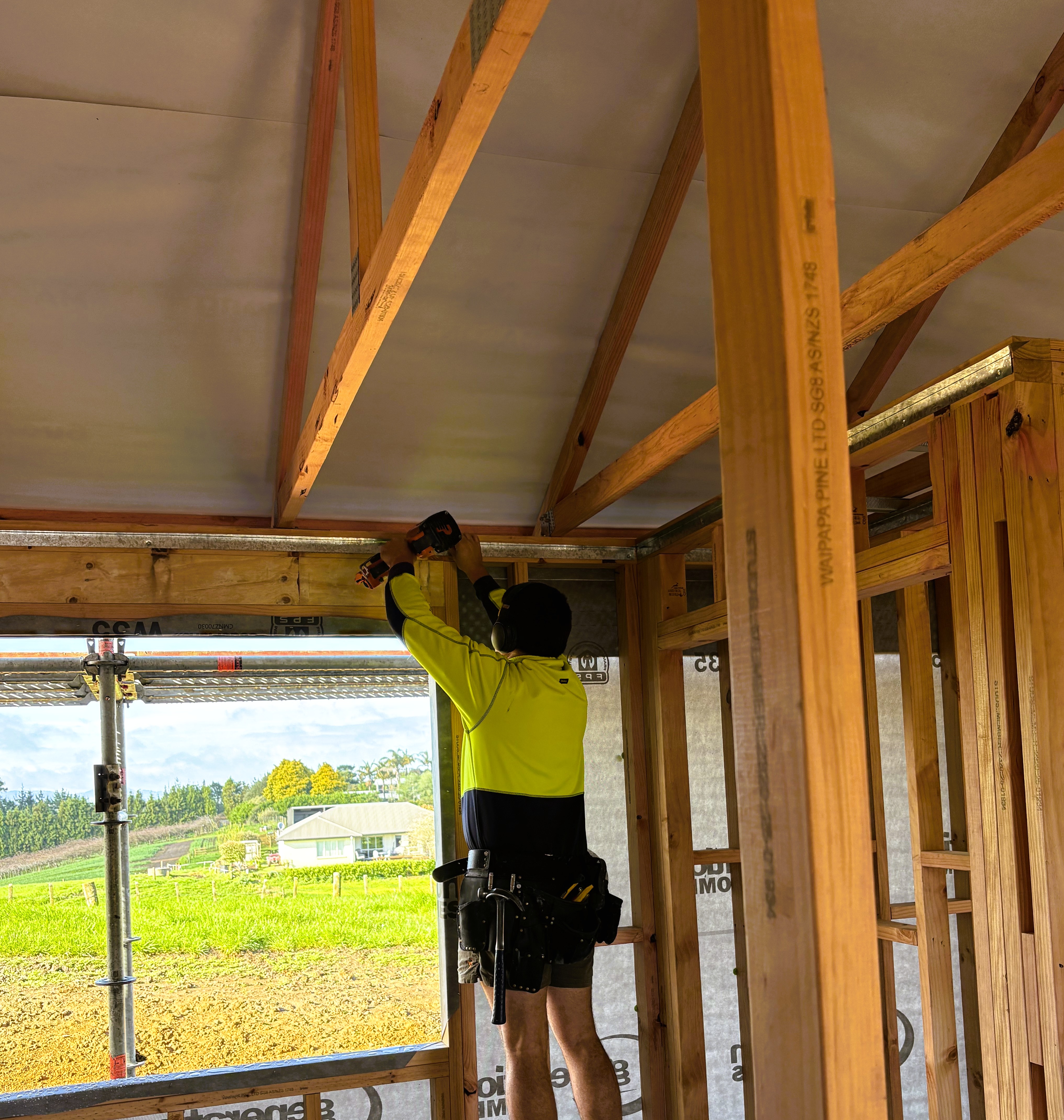 A construction worker in a neon yellow jacket uses a drill while installing metal beams in a house under construction.