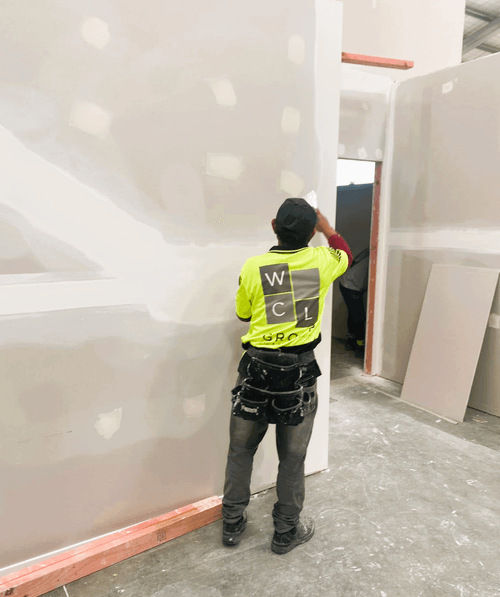 A man in a black T-shirt applies plaster to a wall in a brightly lit room.