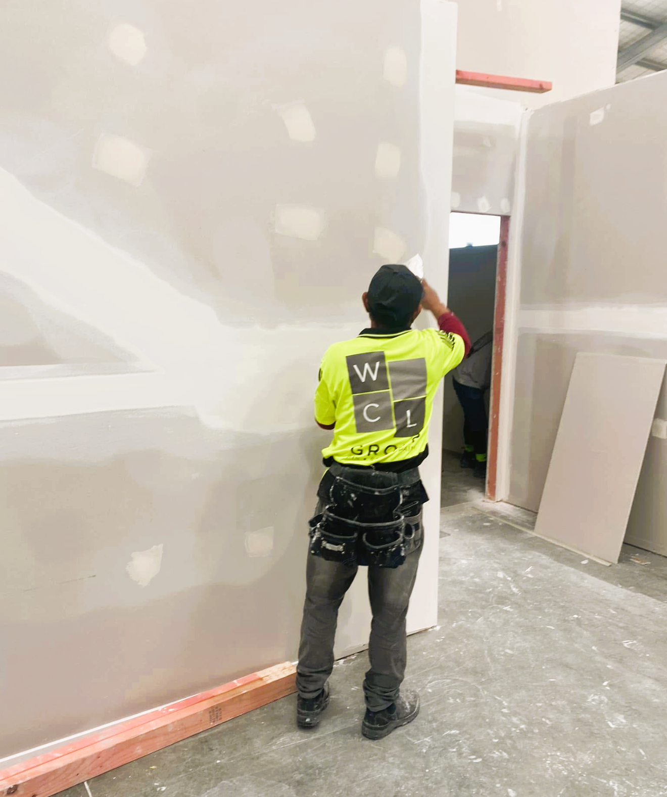 A man in a black T-shirt applies plaster to a wall in a brightly lit room. 
