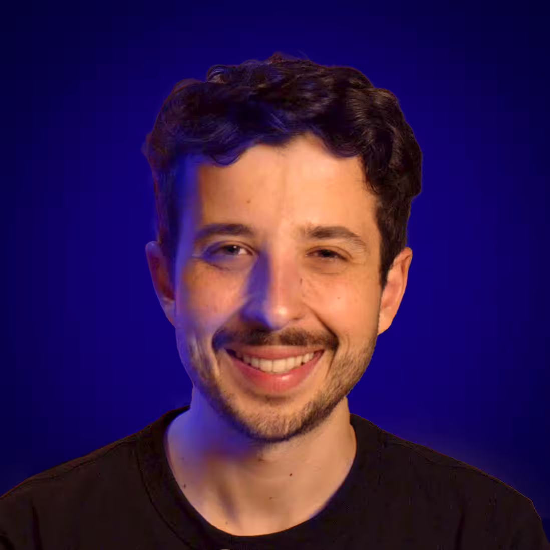 Un homme en tshirt noir, sourit, avec des cheveux noir et un mur bleu en fond