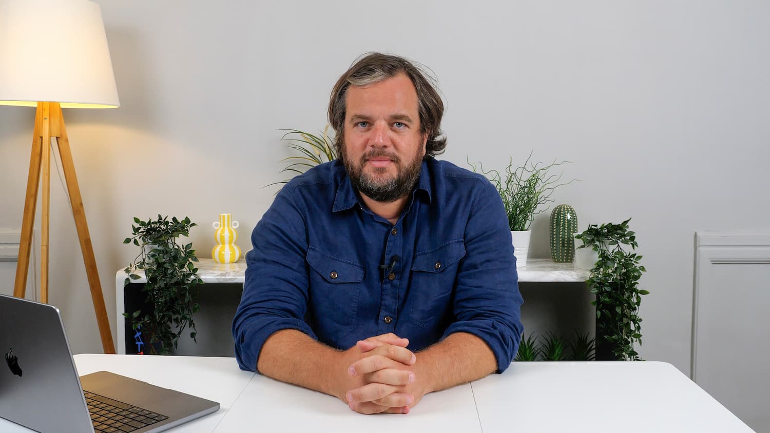 Une photo d'un homme assis à une table. L'homme a une chemise bleue foncée, le mur en arrière est blanc, on peut voir aussi une lampe, un ordinateur et des plantes vertes.