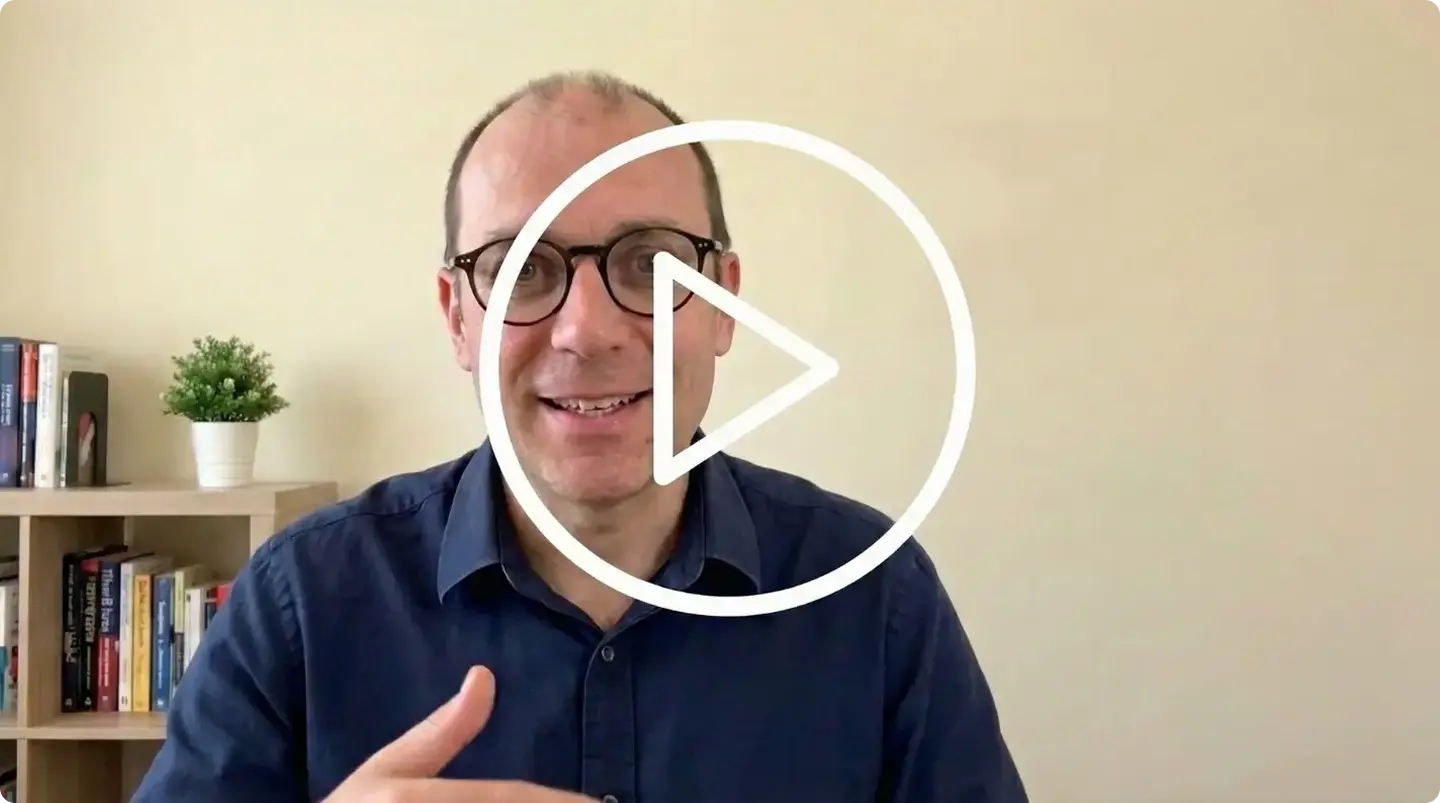 Homme portant des lunettes et une chemise bleue foncée, souriant devant un mur beige avec une étagère contenant des livres et une plante.