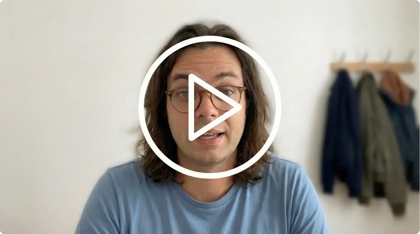 Homme aux cheveux longs et lunettes, portant un t-shirt bleu, posant devant un mur clair avec des manteaux accrochés à un porte-manteau.