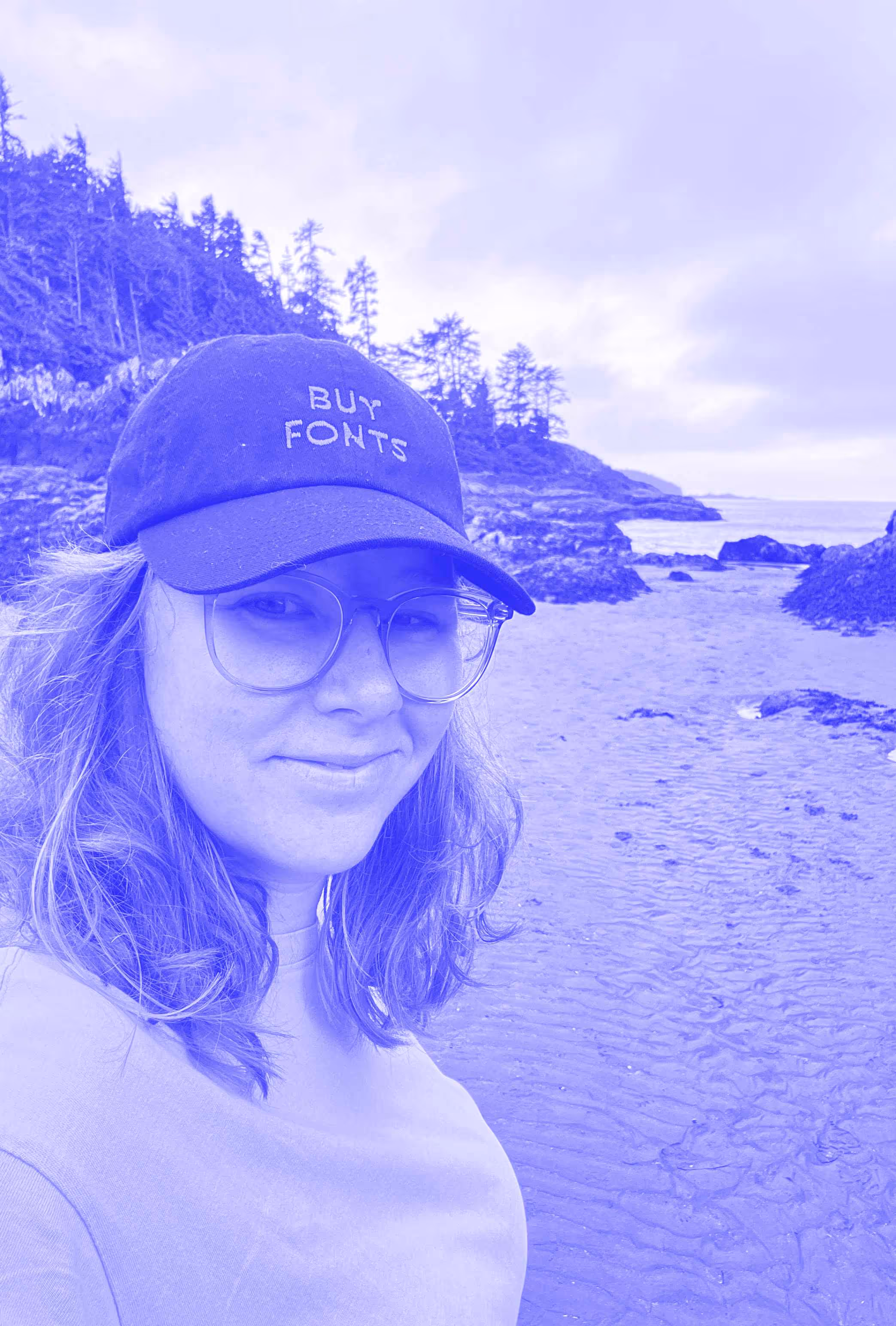 A photo of Alanna with a west coast beach in the background. She is wearing a hat that says, "buy fonts."