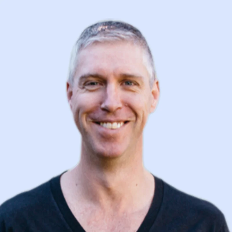 Smiling man with short grey hair wearing a black v-neck t-shirt, on a light blue background.
