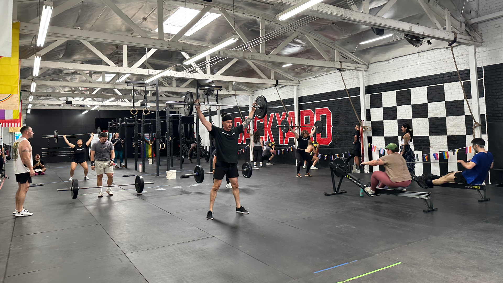 A busy gym with people weightlifting, rowing, and working out. The industrial space has white beams and checkered walls, creating an energetic atmosphere.