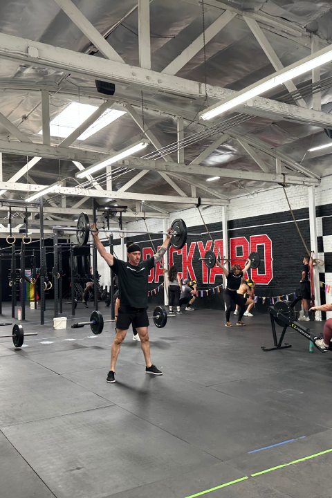 A busy gym with people weightlifting, rowing, and working out. The industrial space has white beams and checkered walls, creating an energetic atmosphere.