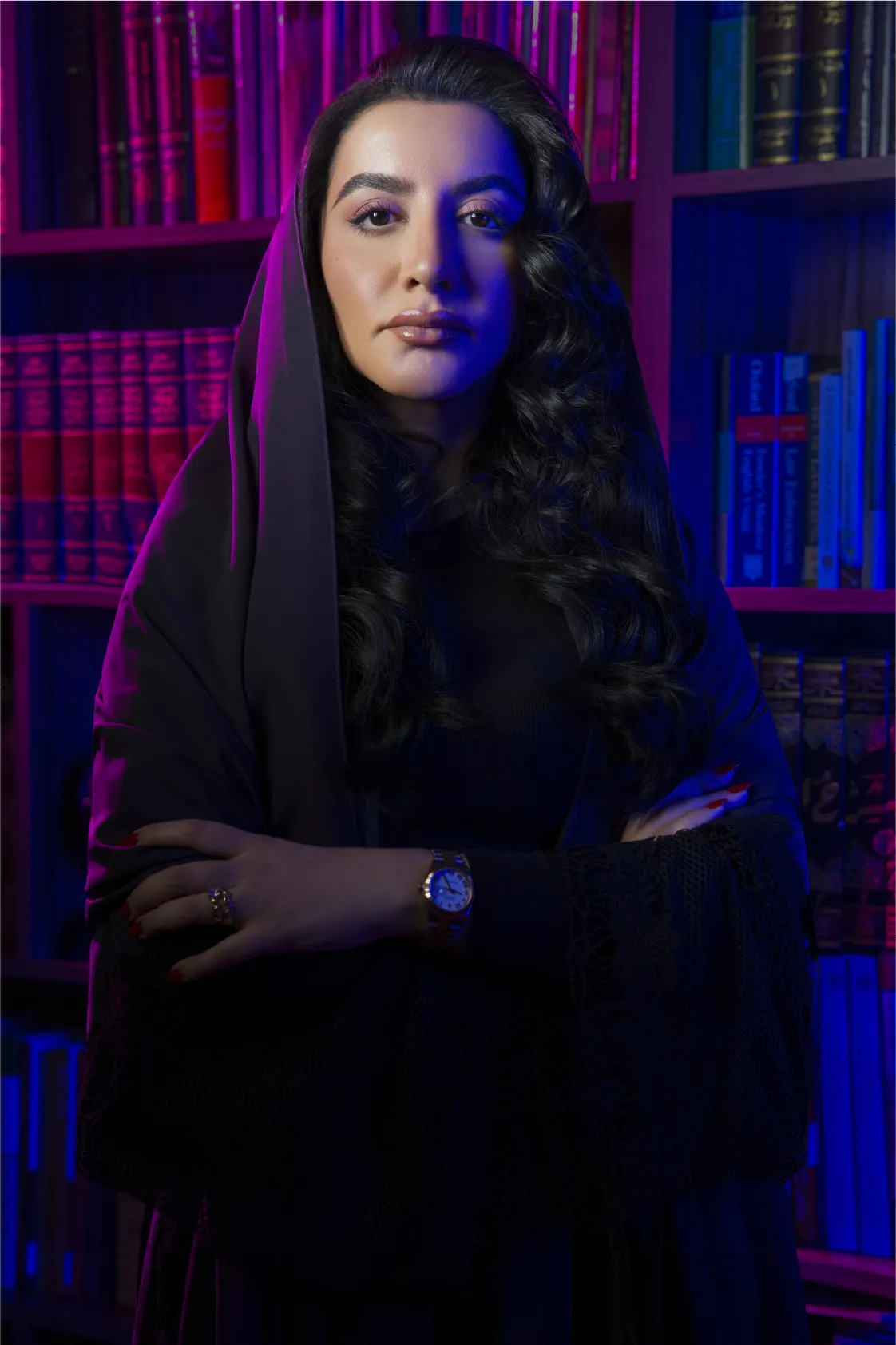 Portrait of a confident woman with long dark hair in a white headscarf and dress, arms crossed against a blue background.