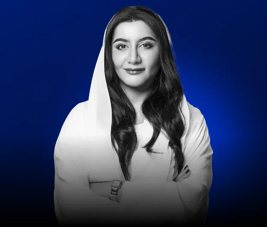 Portrait of a confident woman with long dark hair in a white headscarf and dress, arms crossed against a blue background.