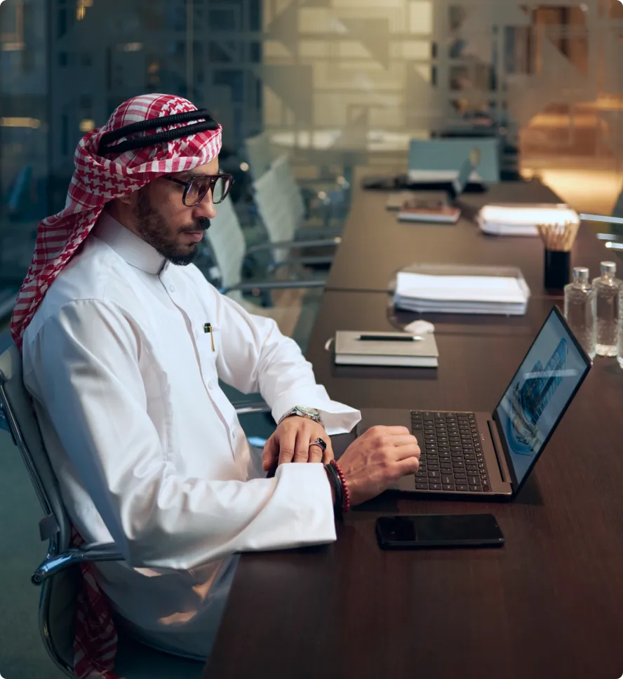 Smiling woman wearing a light gray hijab and round glasses working on a laptop in a modern office.