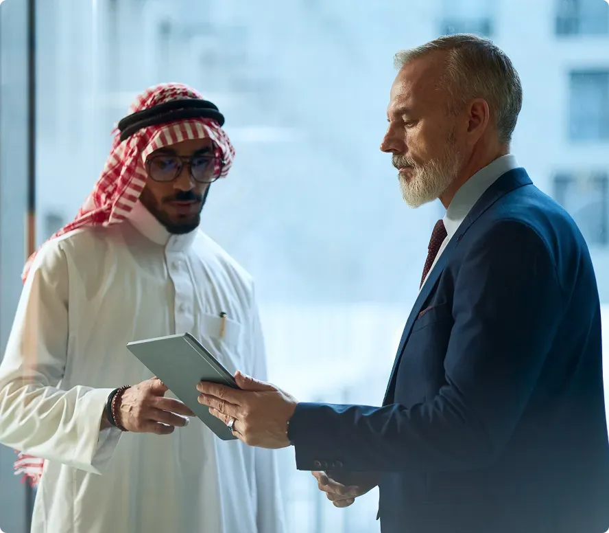Two businessmen, one in traditional Middle Eastern attire and the other in a blue suit, looking at a tablet together.