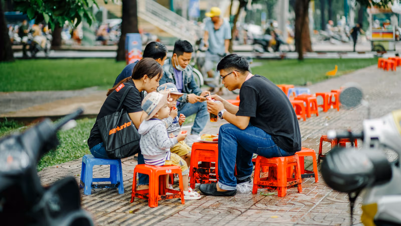 A family with two children sitting on small red and blue stools at an outdoor area, eating and drinking together.
