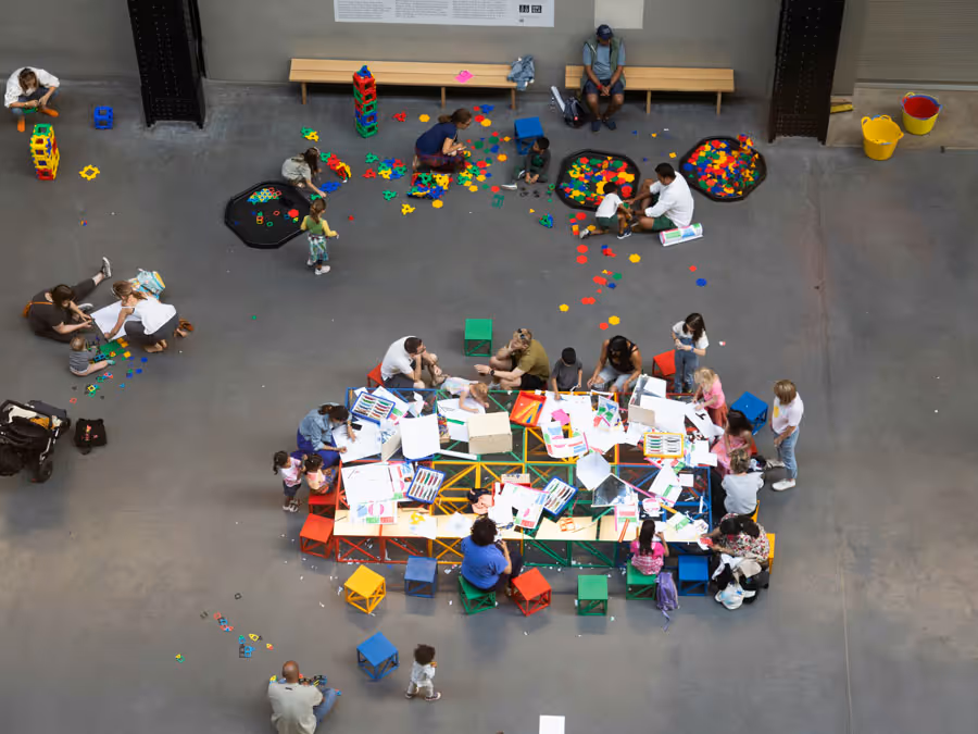 Overhead view of children and adults seated around a large table with art supplies, surrounded by scattered colorful building blocks on a concrete floor.