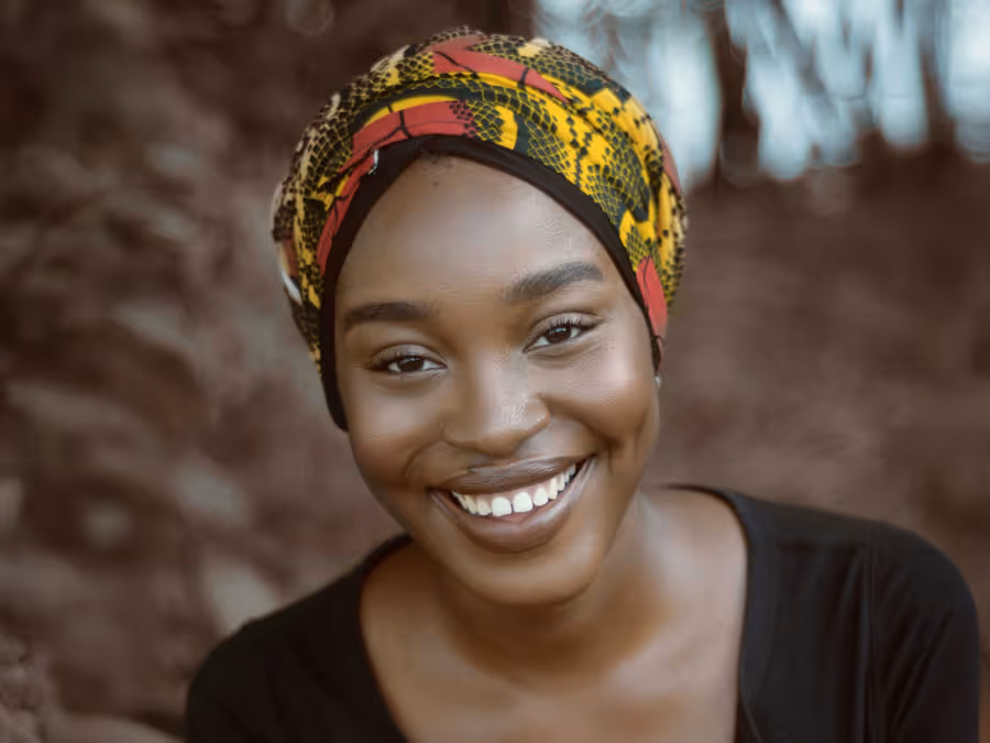 Smiling woman wearing a colorful yellow and red patterned headscarf and black top, with a blurred natural background.