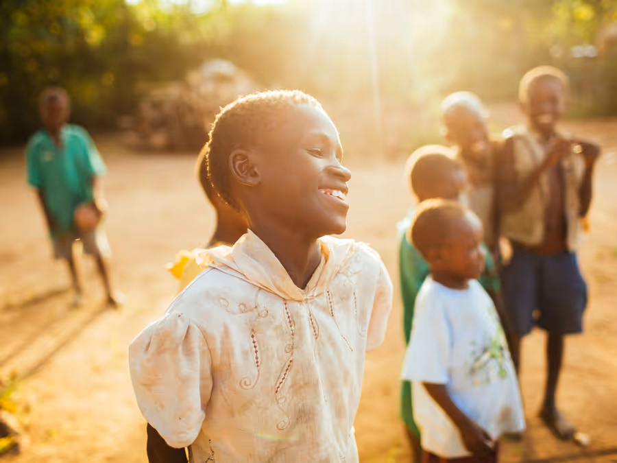 Smiling child in a light-colored hoodie with other children and warm sunlight in the background.