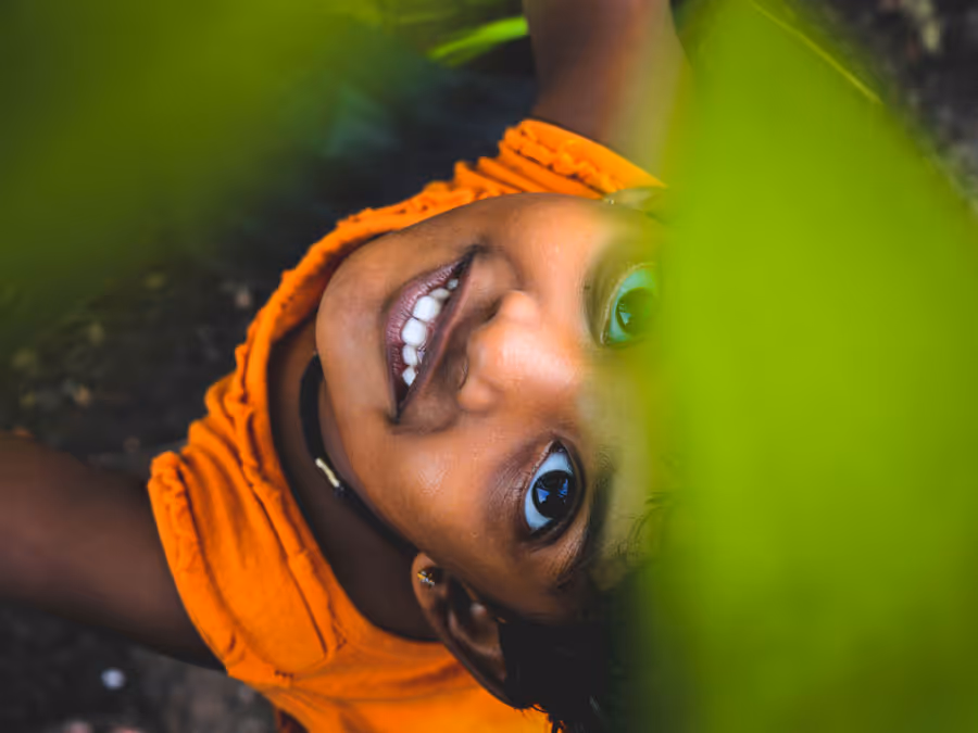 Smiling child in an orange top looking up through green leaves.