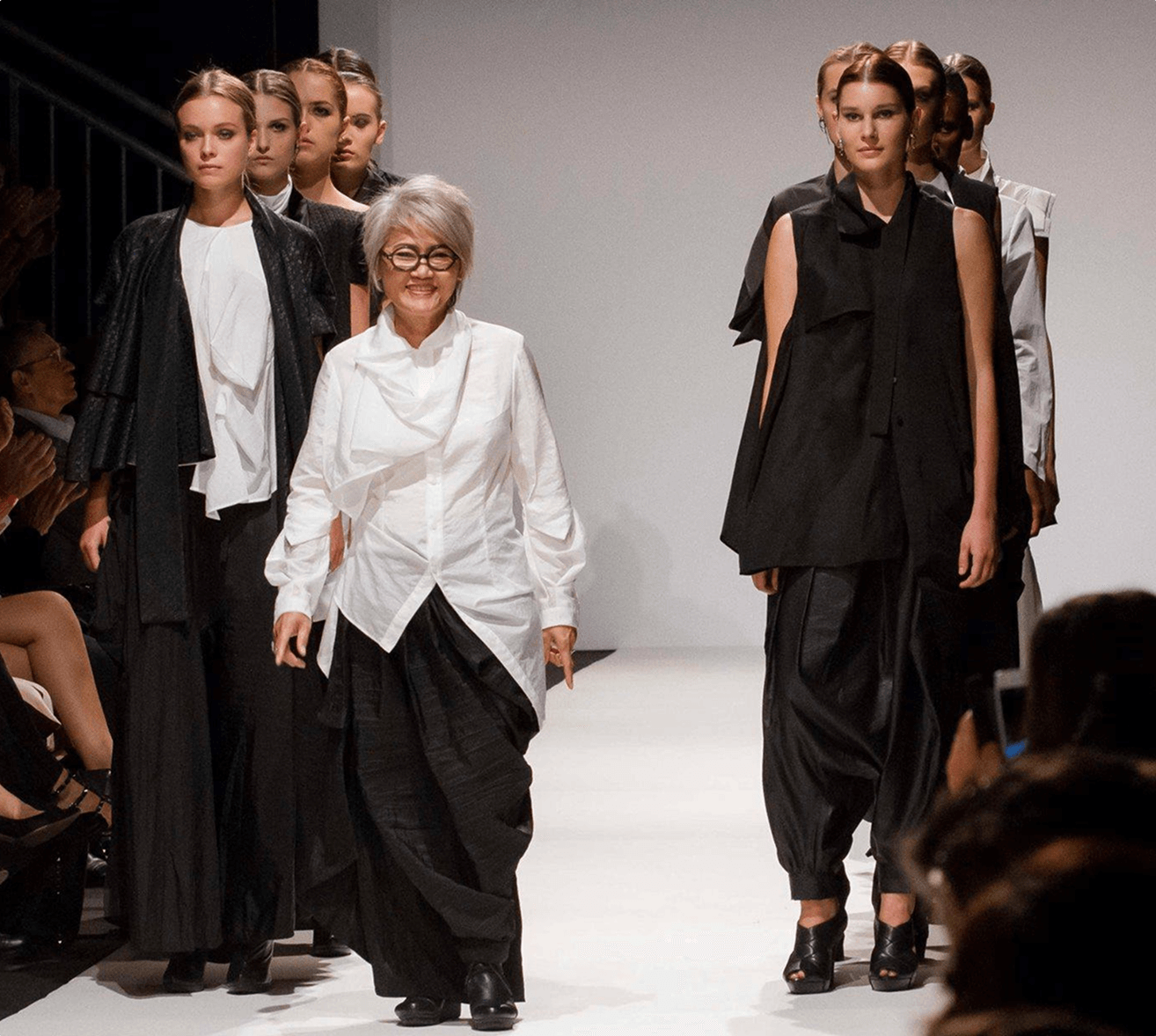 Fashion designer walking on a runway with models in black and white avant-garde clothing behind her.