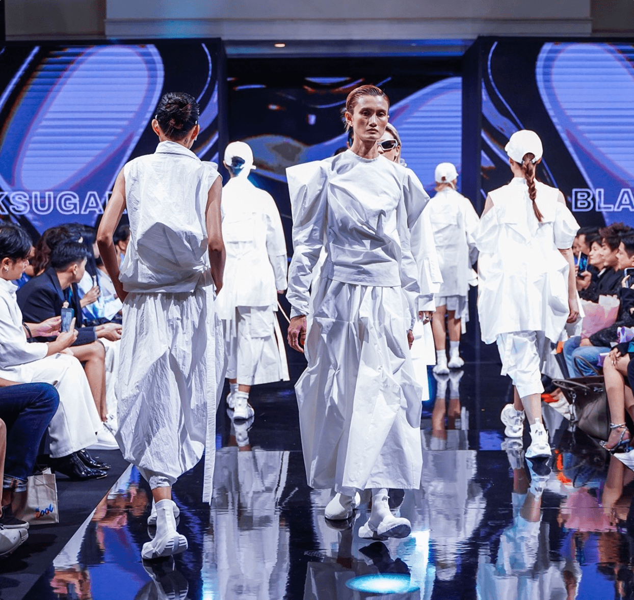 Models walking a runway in futuristic, all-white outfits during a fashion show with an audience seated on both sides.